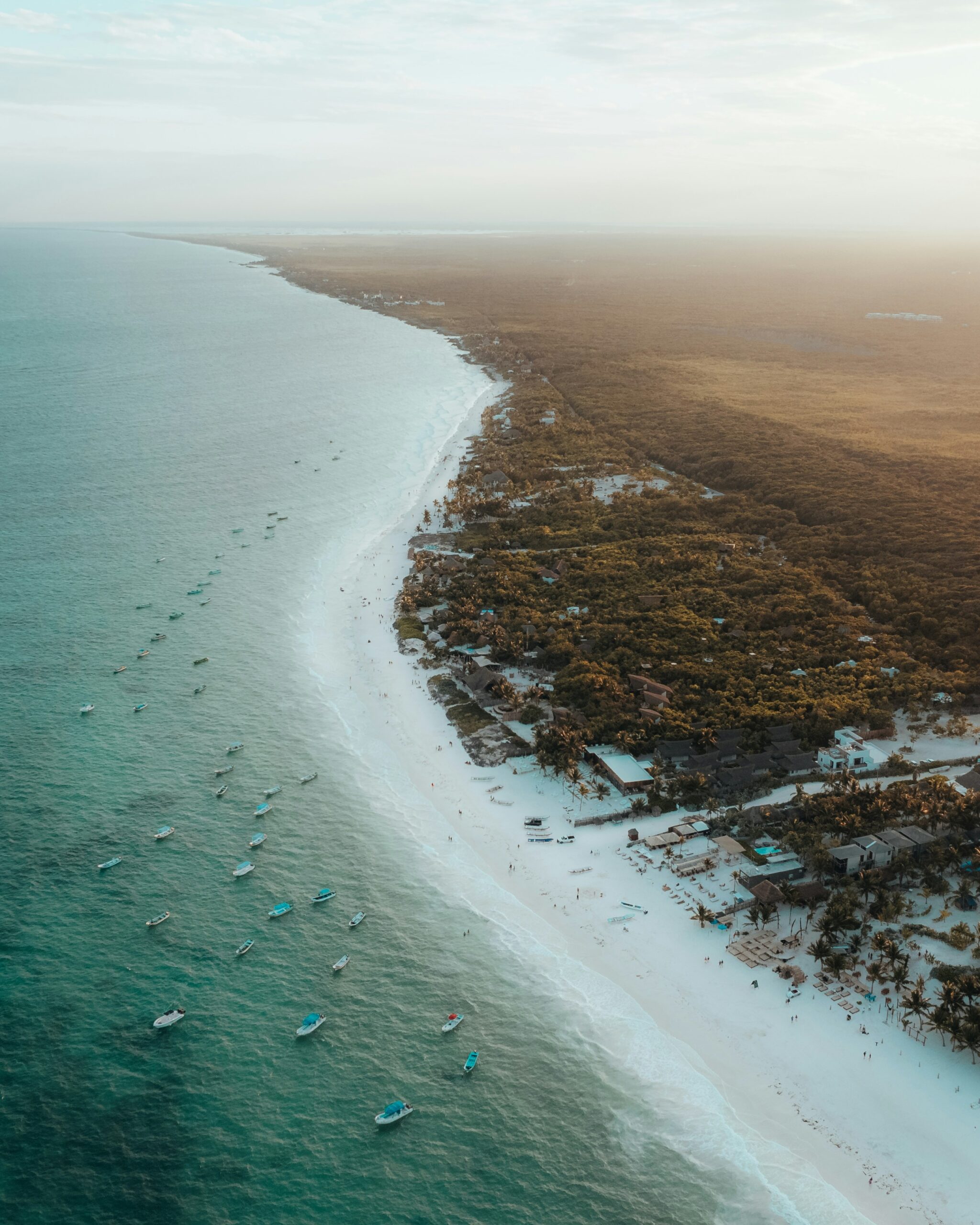 Aerial view of a white-sand beach in Mexico with turquoise water, small boats offshore, and beachfront homes surrounded by palm trees.