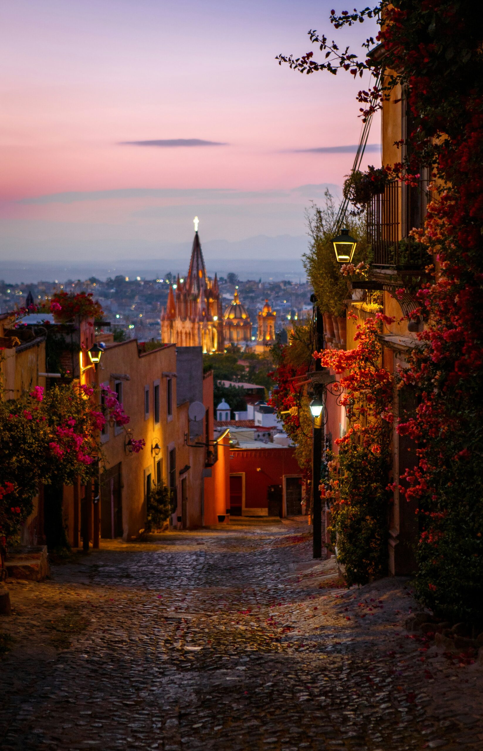 Cobbled street in a Mexican colonial town at sunset, lined with colorful buildings, blooming bougainvillea, and warm lantern light, with a cathedral illuminated in the distance.
