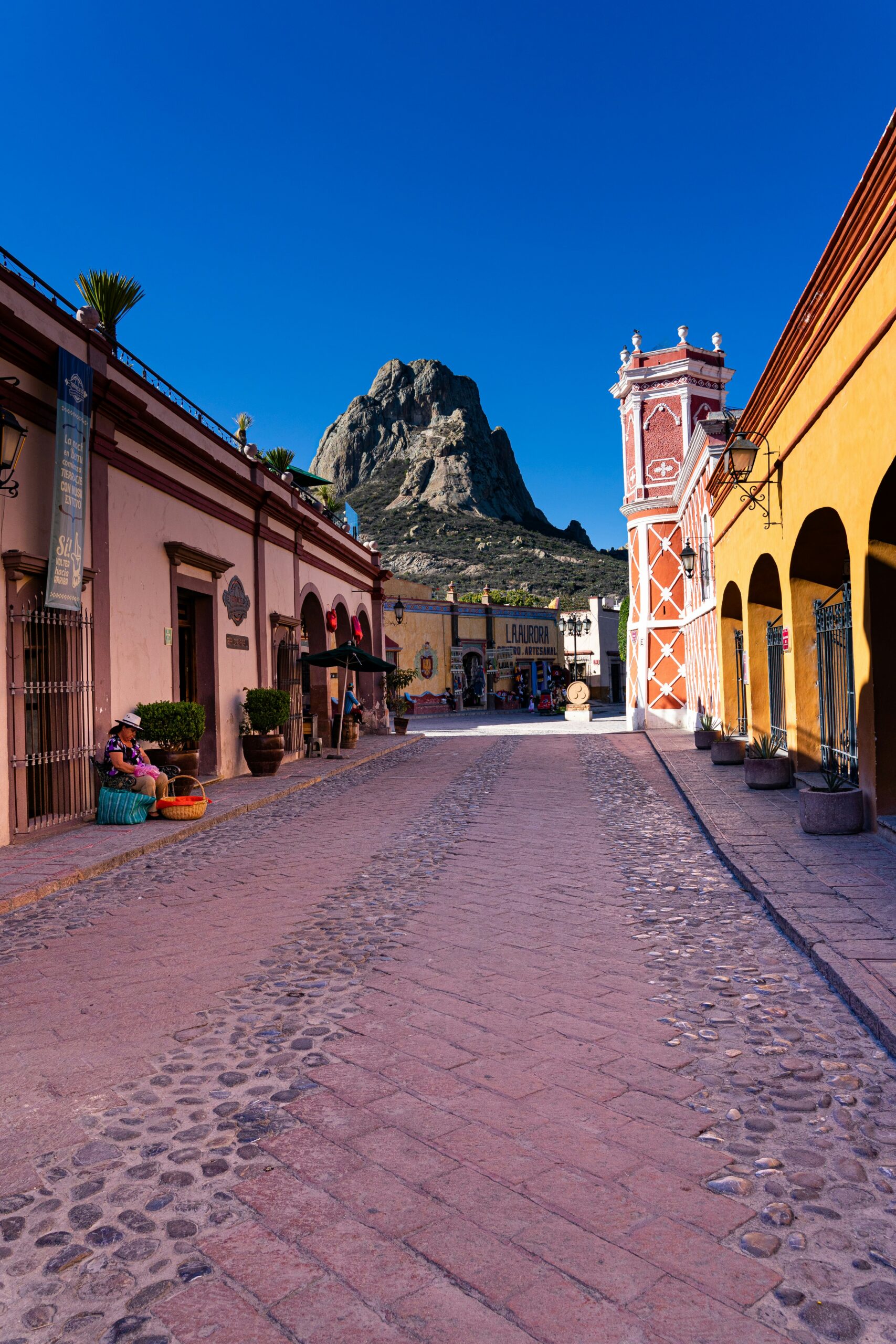 Cobbled street in a colorful Mexican town with pastel buildings, arched walkways, and a mountain rising in the background under a clear blue sky.